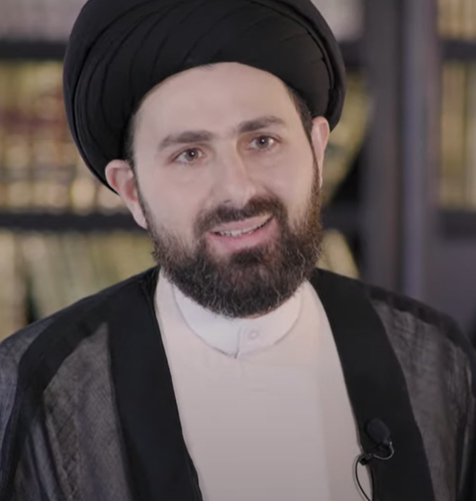 Image of a smiling man with a beard and black turban, sitting in front of a blurred library. 