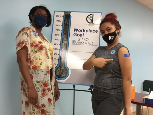 Two people wearing masks pose in front of a vaccine tracker poster while one shows a vaccine bandage on their arm