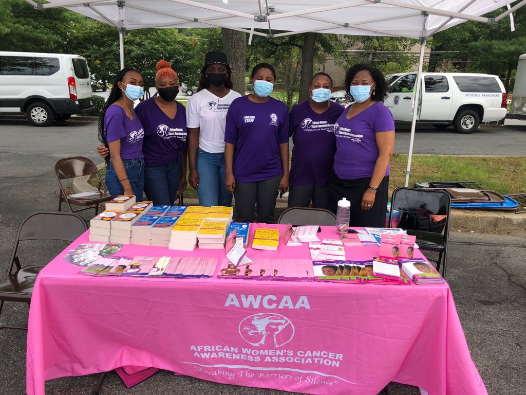 A group of masked volunteers pose together behind a table that contains literature and books at an outdoor event,