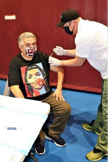 A man wearing a mask sits in a chair and receives a COVID vaccine from a man wearing a black hat