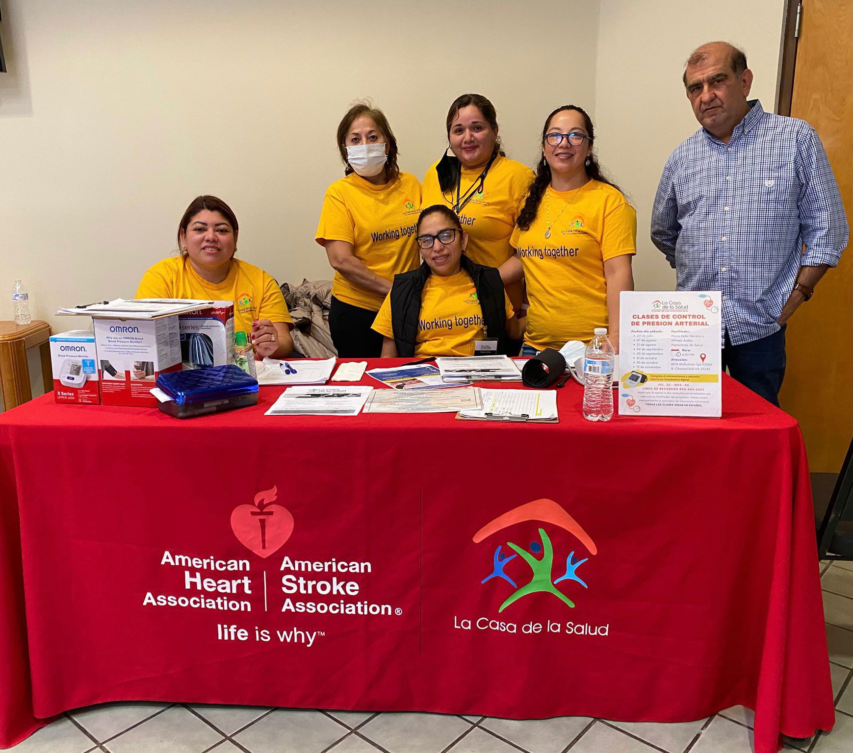 A group of volunteers smiles and poses together at a table. The table cloth notes they are there with the American Heart Association, American Stroke Association, and la Casa de la Salud