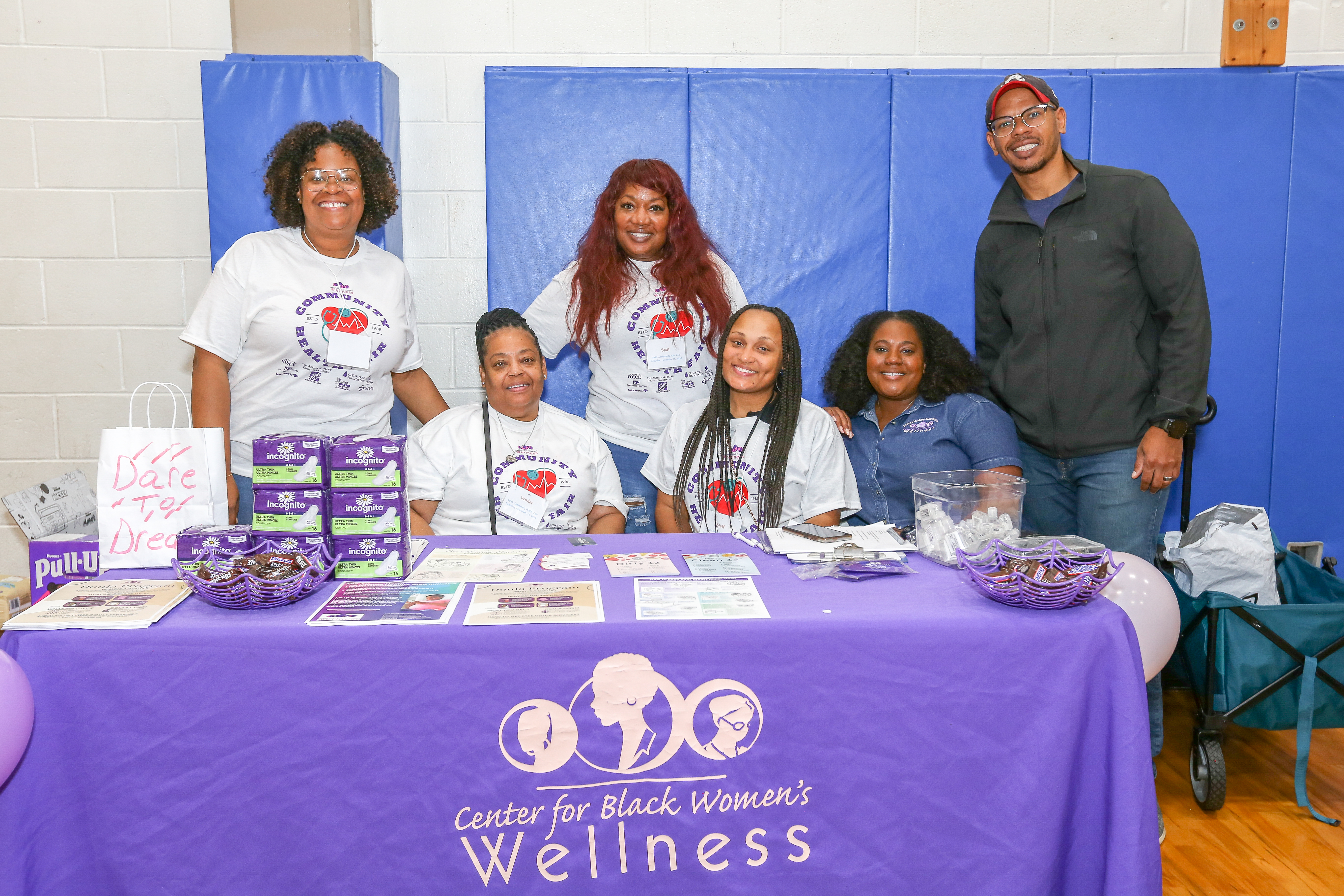 A group of young Black women and one Black man gather around a table with a banner that says "Center for Black Women's Wellness"