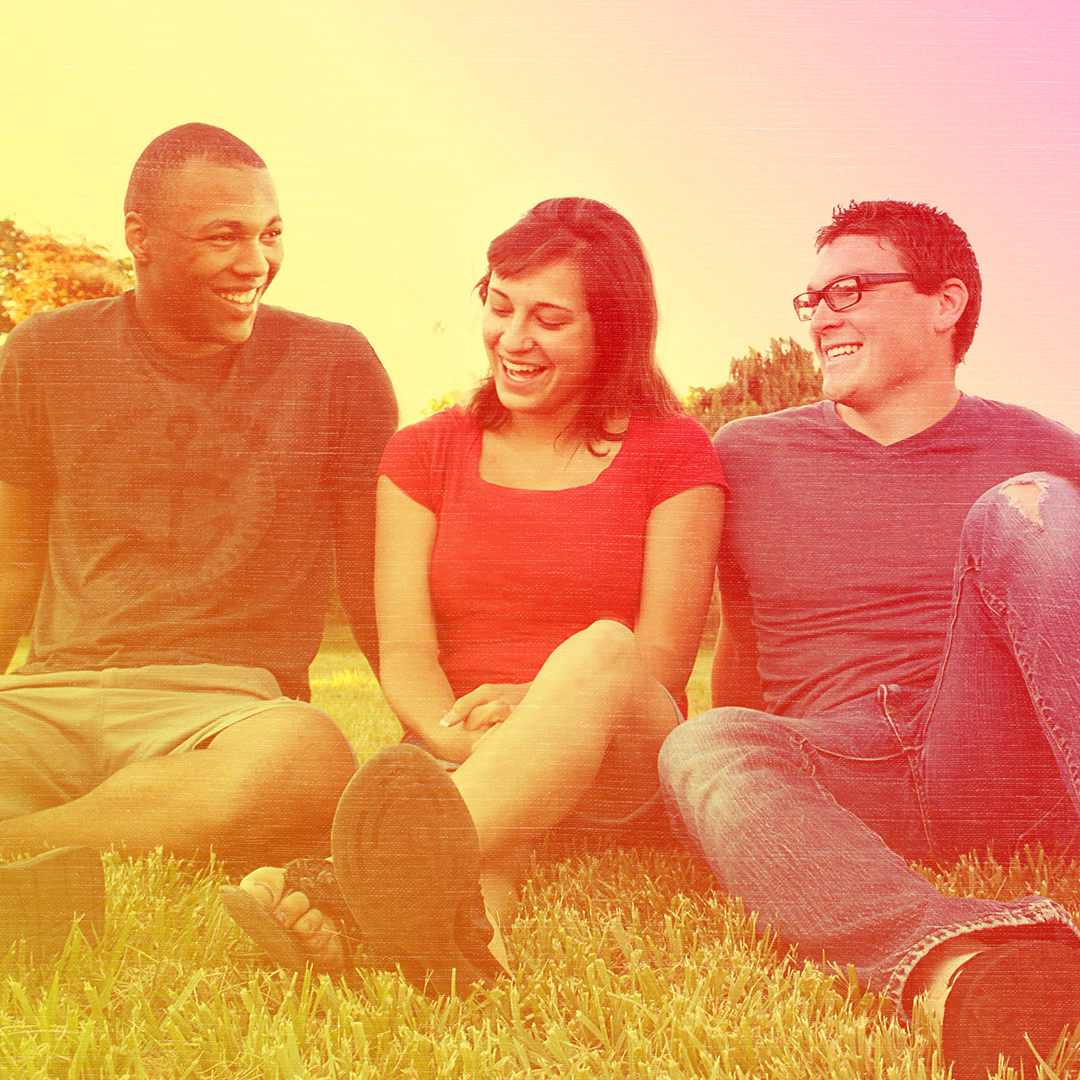Red colorized photo of three people sitting on the grass smiling. A white young woman sits between a Black young man and white young man.
