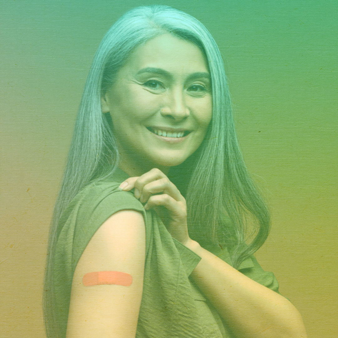 greenish-yellow filtered photo of a woman with long gray hair smiling and showing her arm with a bandage from receiving a vaccine