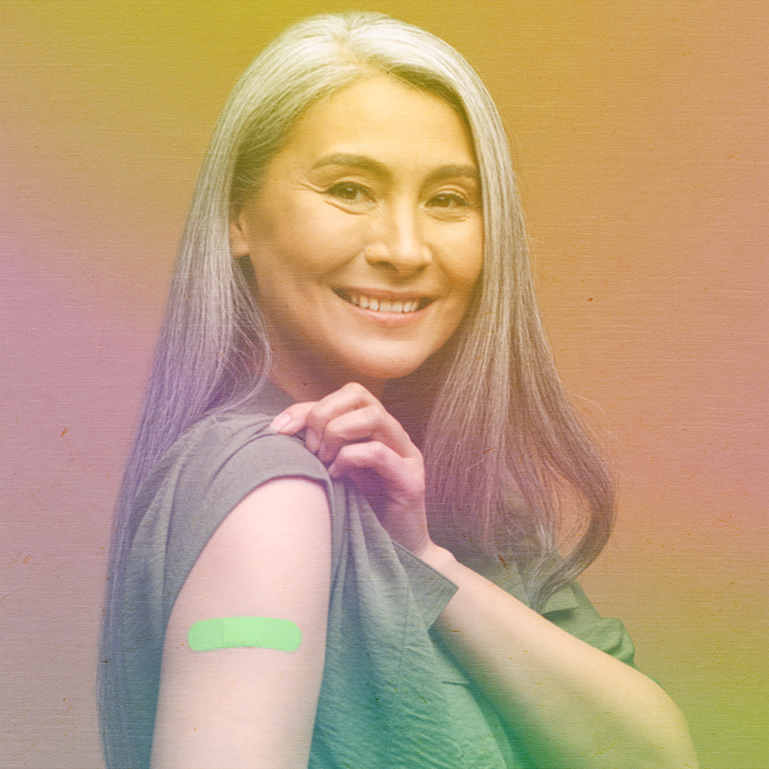 color photo of a woman with long gray hair smiling and showing her arm with a bandage from receiving a vaccine