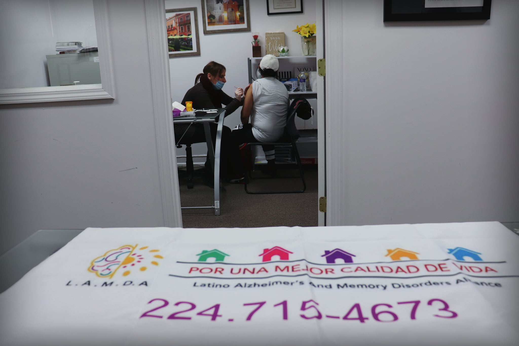 A woman administers a vaccine behind a table with a LAMDA banner