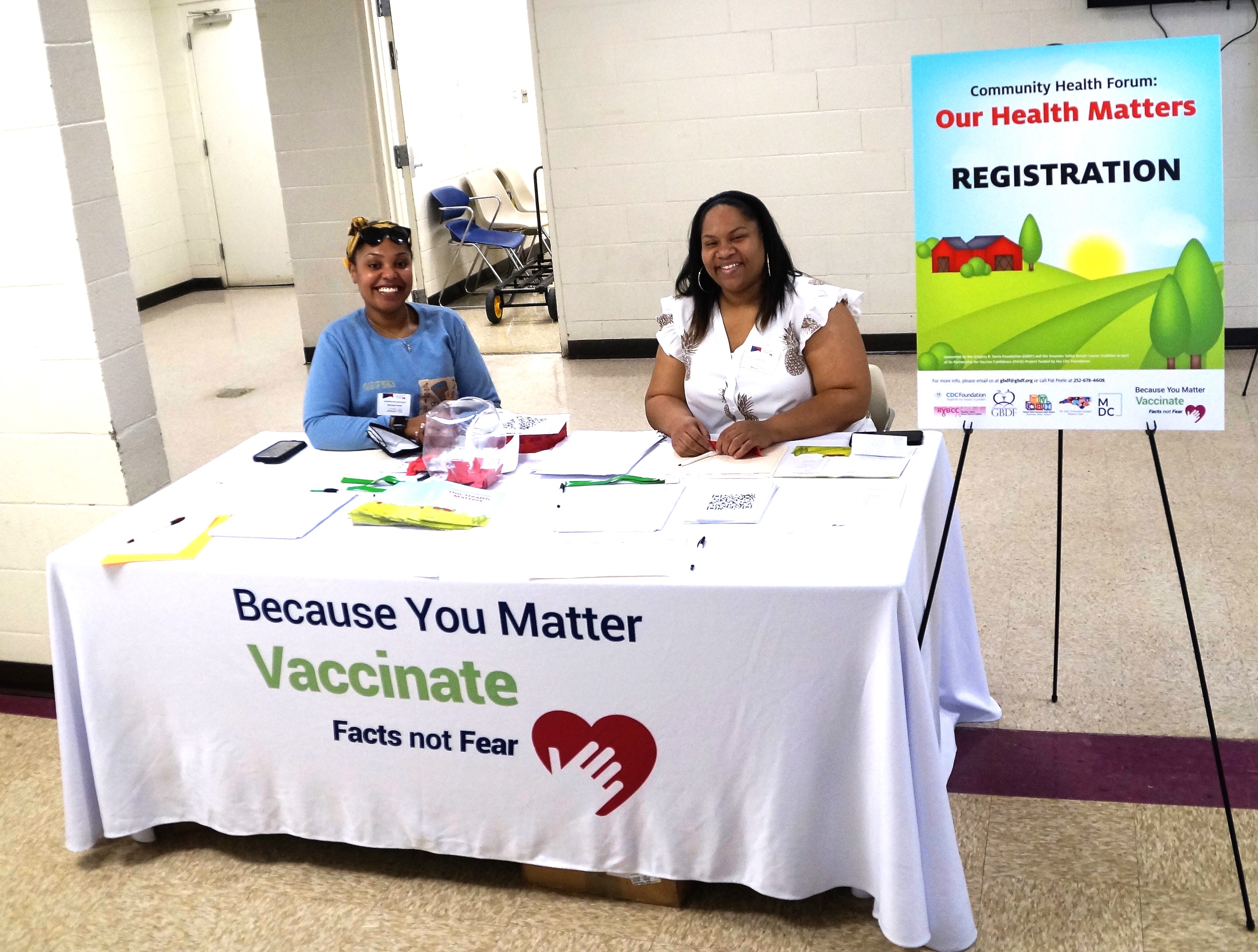 Two Black women sit at an informational table at a community vaccine event. 