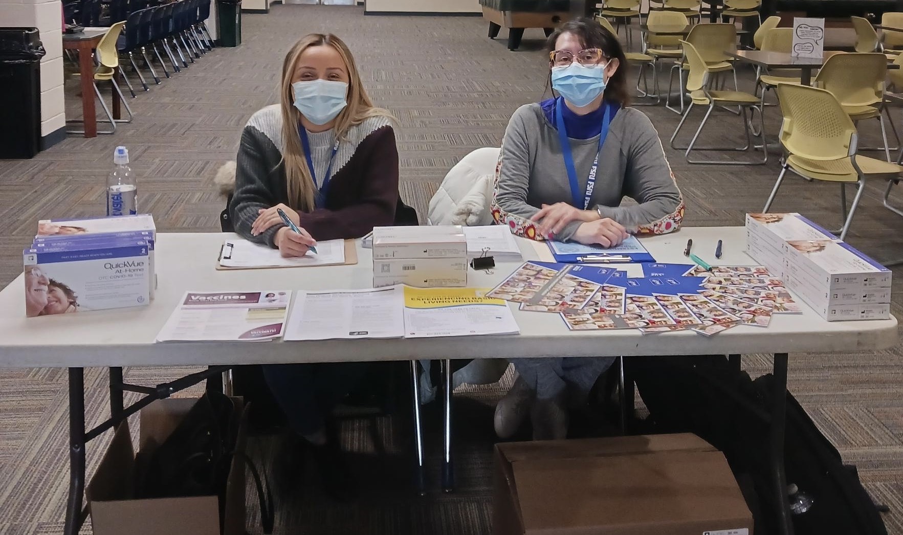 Two women wear masks and sit at a community health event table