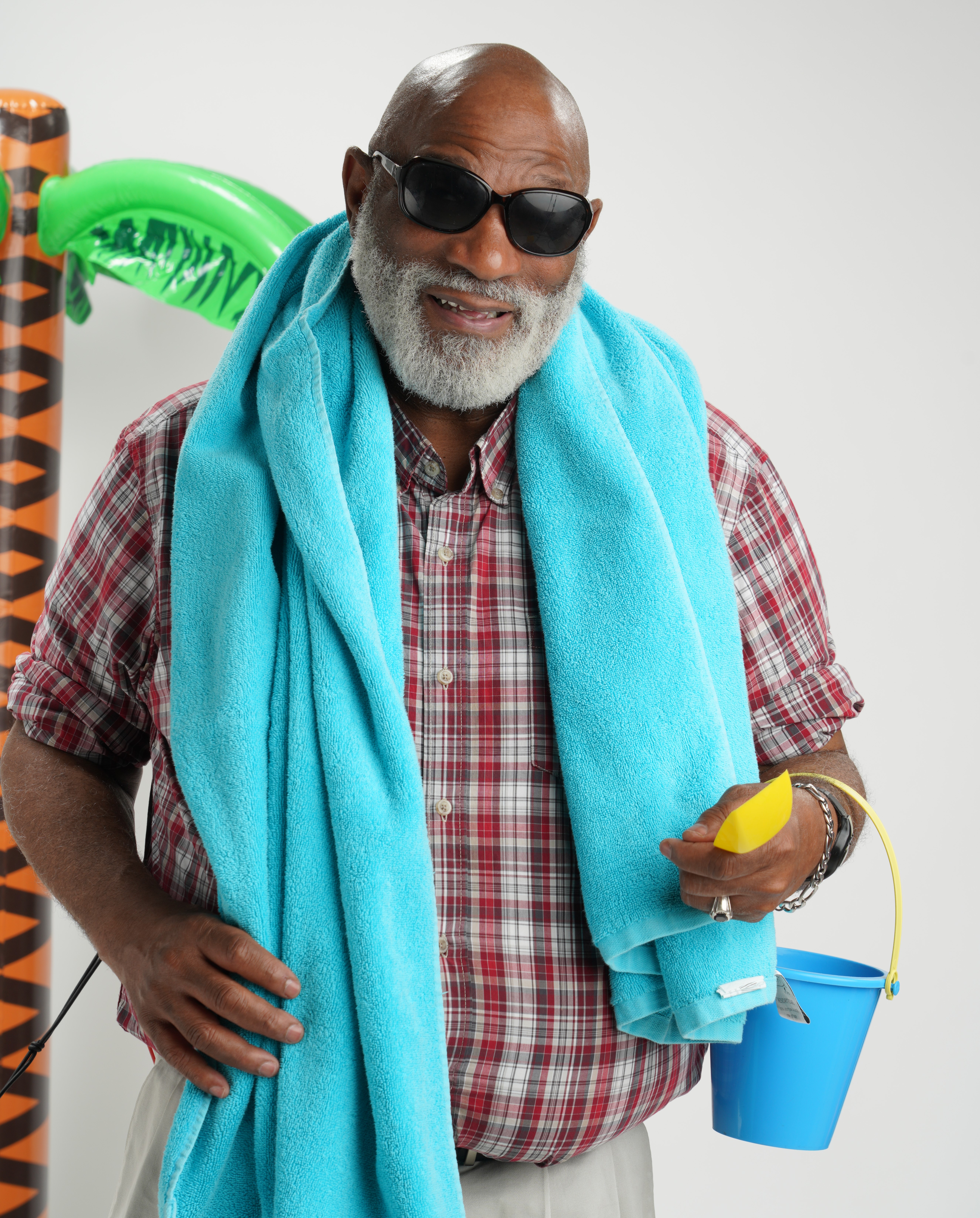 A Black older man wears sunglasses and a beach towel around his neck and carries a blue bucket. Inflatable palm tree in the background.