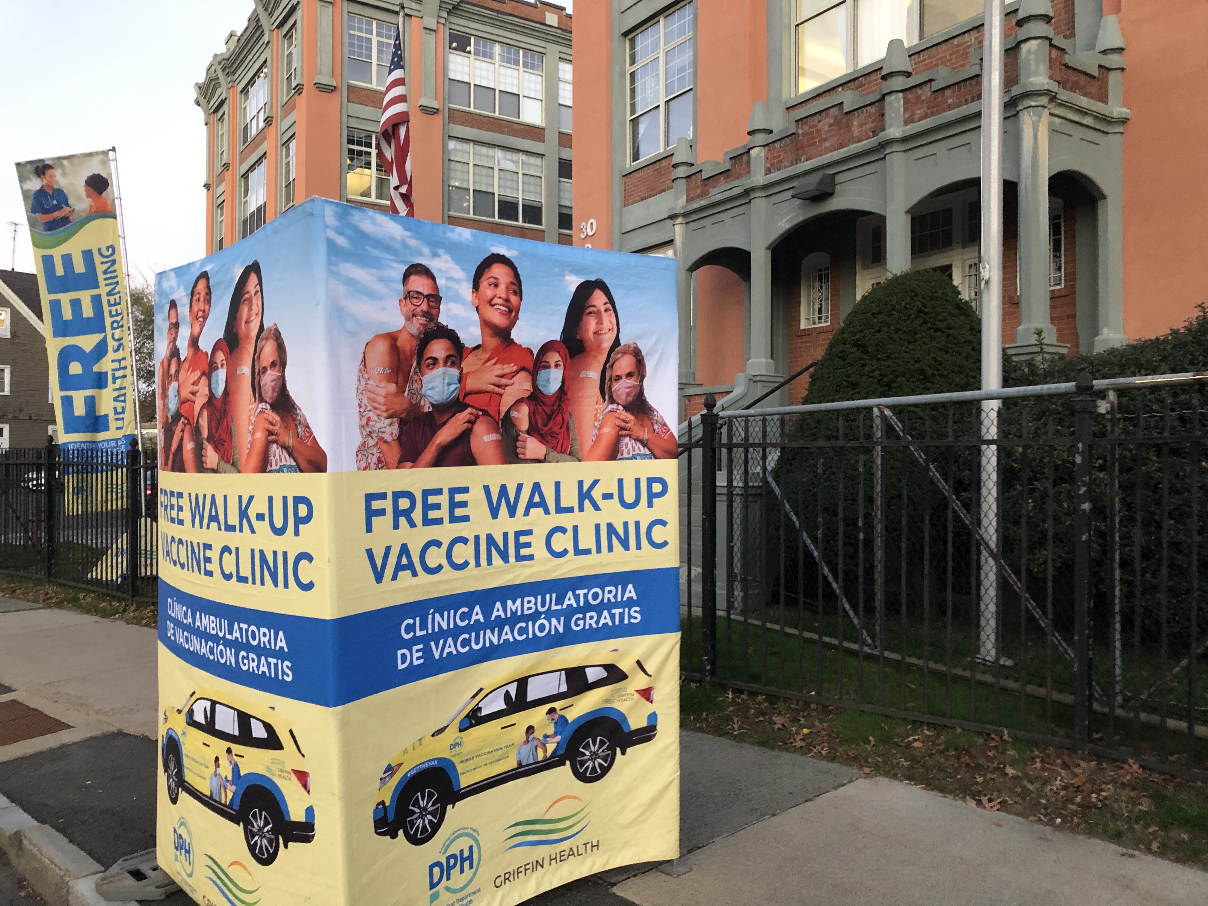 Image shows a sign on the sidewalk for a free walk-up vaccine clinic, with the text in both English and Spanish.
