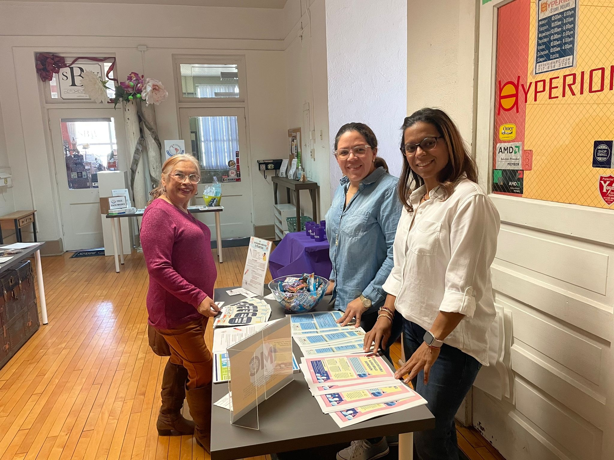 A group of three women of color smile and stand around a table displaying educational materials about vaccines.