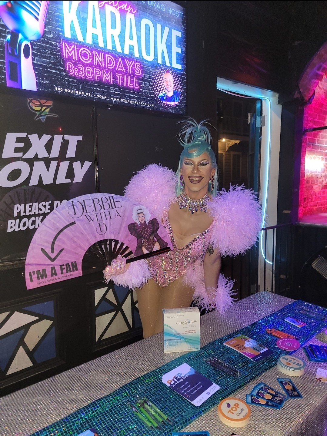 Drag queen stands behind a bedazzled resource table, holding a big pink fan and wearing a pink feather leotard.