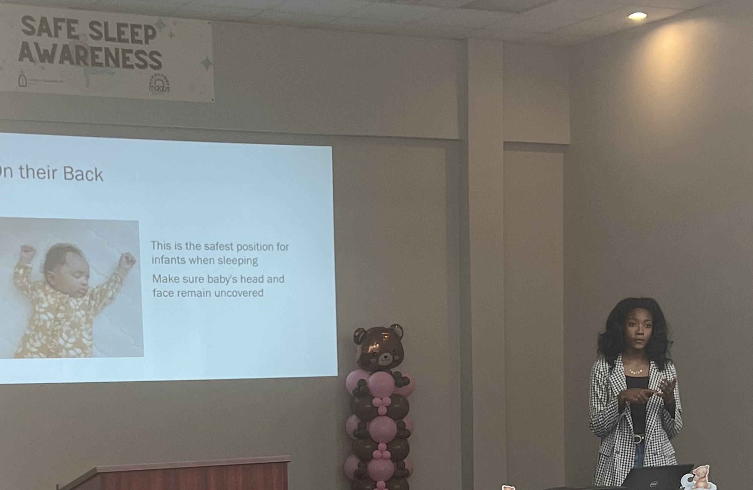 A Black woman stands behind a table and gives a presentation, with a PowerPoint projected behind her showing a picture of a Black newborn.