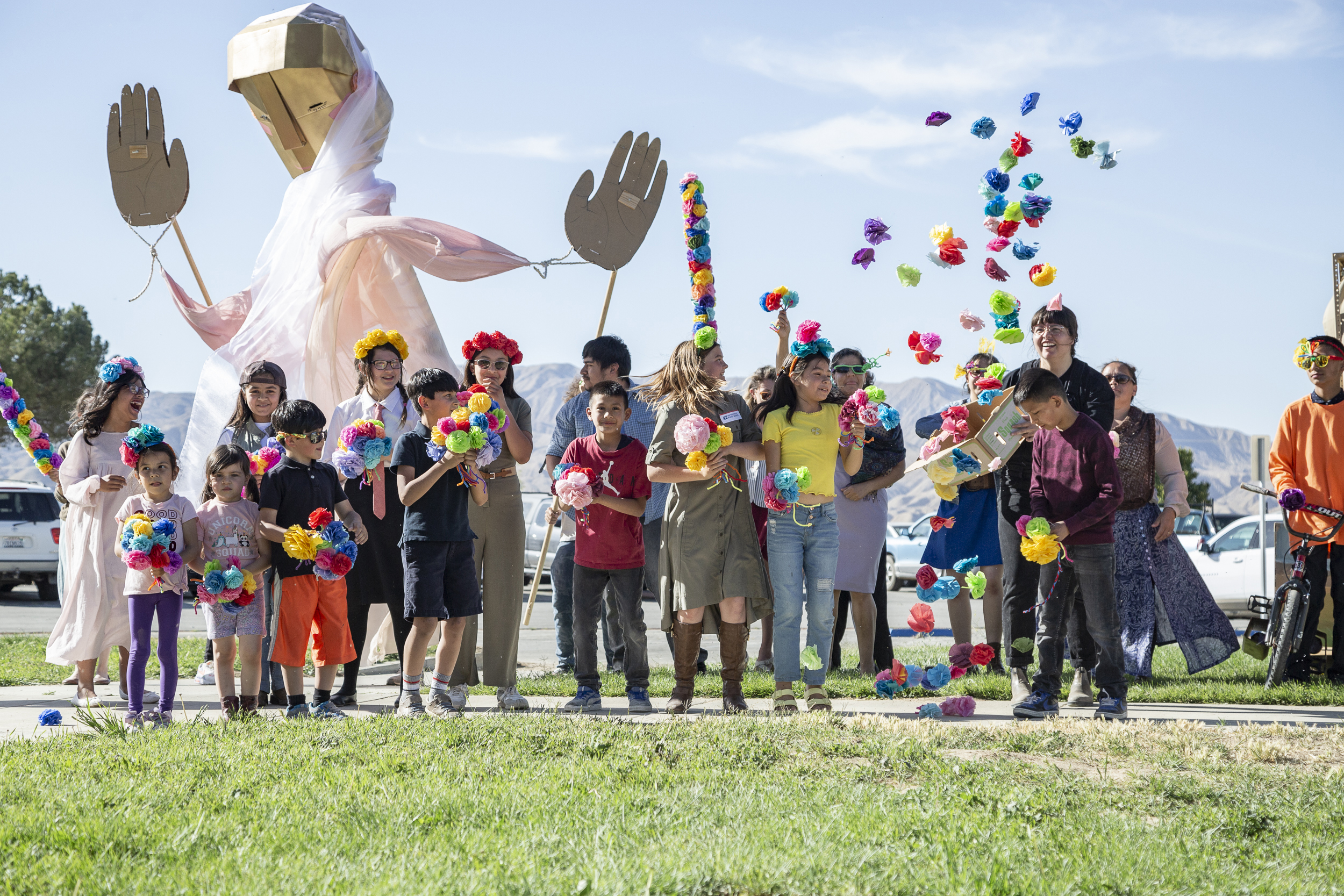A large group of smiling children stand in a line on the sidewalk and throw paper flowers in the air.