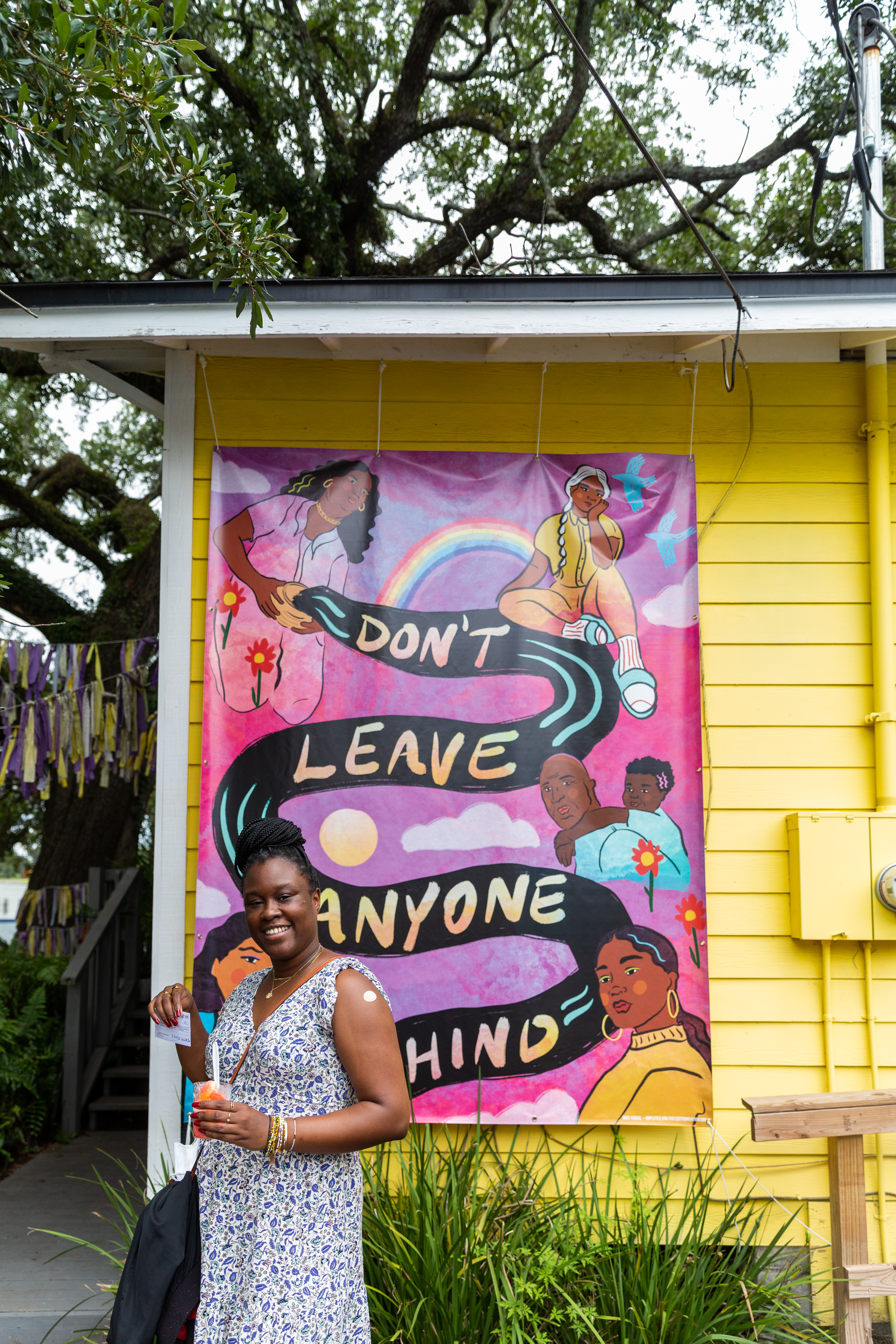 A Black woman smiles and holds up her COVID-19 vaccine card in front of a yellow building with a sign that reads "don't leave anyone behind."