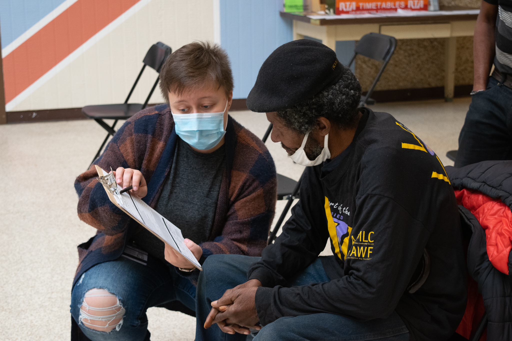 A person wearing a mask reads text on a clipboard to a Black man 