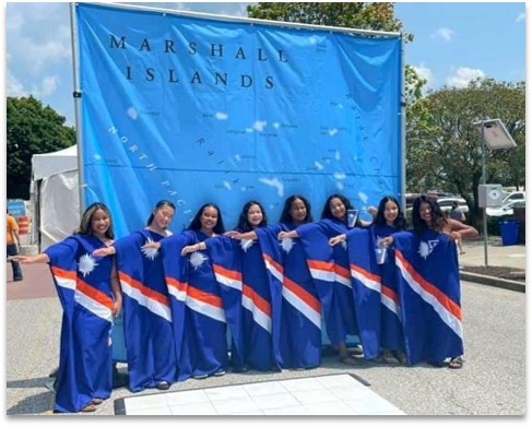 Marshallese women in traditional attire stand in front of a map of the Marshall Islands.