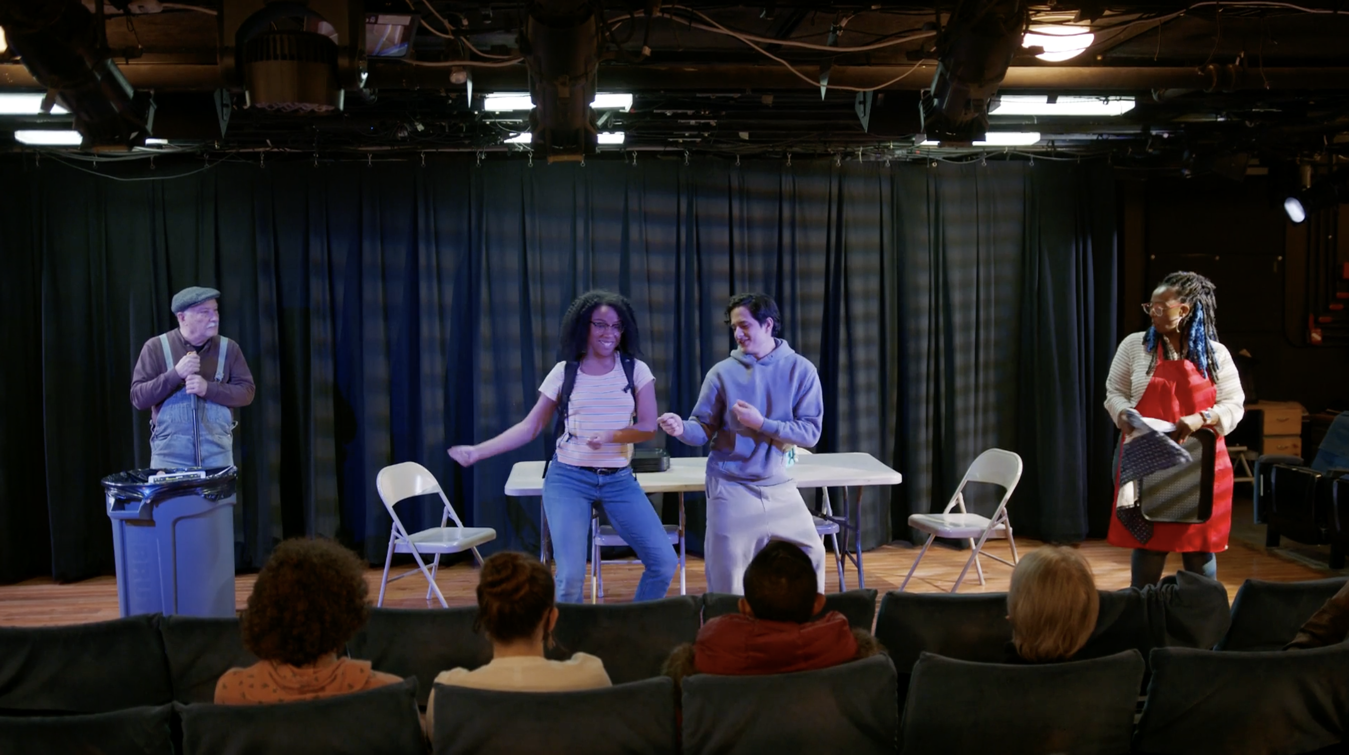 Actors smile and dance on stage in front of a folding table and chairs.