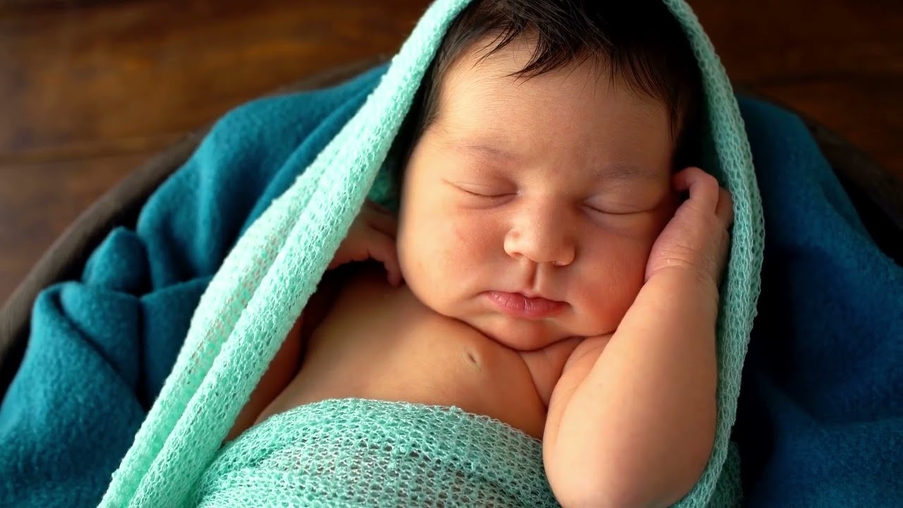 Image of a  newborn with dark brown hair sleeping with a blue blanket wrapped around them.
