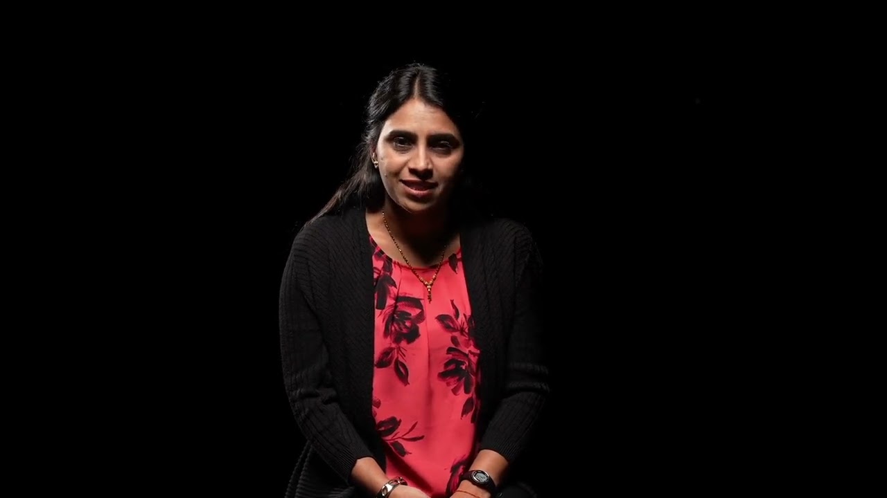 An Indian woman wearing a black sweater and red shirt is speaking. 