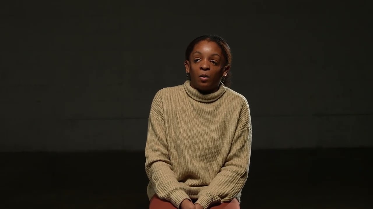 A young Black woman wearing a sweater is speaking. 
