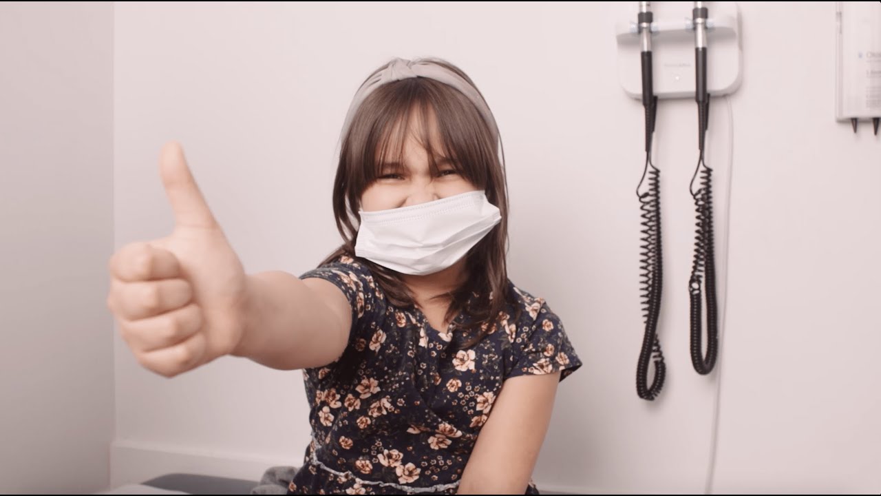 American Indian young girl with a mask and giving a thumbs up while sitting in an exam room.