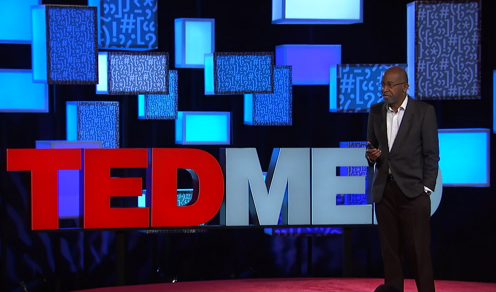 David R. Williams, a Black man, stands on a red carpet in front of a TEDMED screen. He is wearing a black suit, holding a clicker, and speaking into a microphone. 