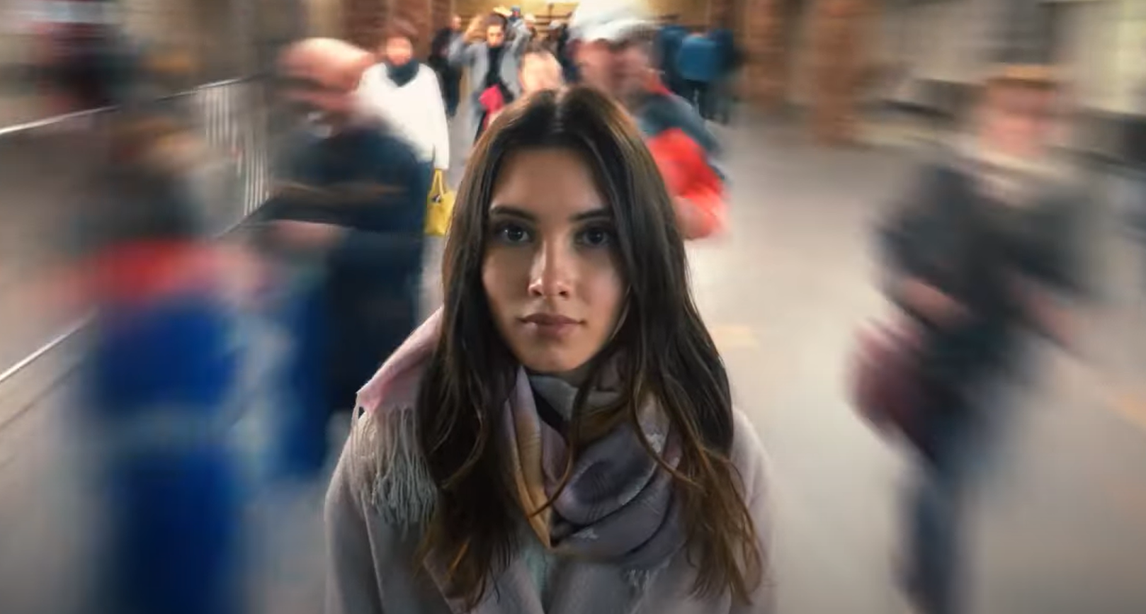 Person with long hair stands still and stares wide-eyed at the camera in a crowded public place, with blurry people buzzing around them.