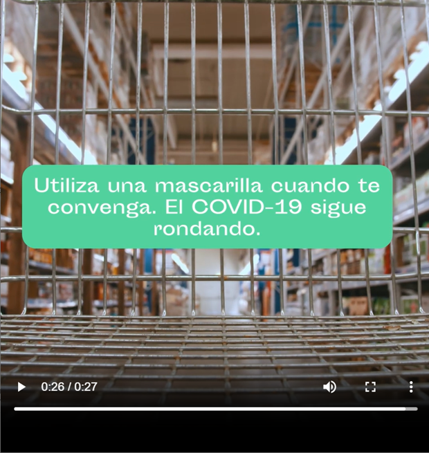 Screenshot of cart in grocery aisle. Text reads in Spanish ' Mask up when it works for you, COVID-19 is still out there.' Example reasons to mask up in video include being immunocompromised and not wanting to catch COVID on vacation.