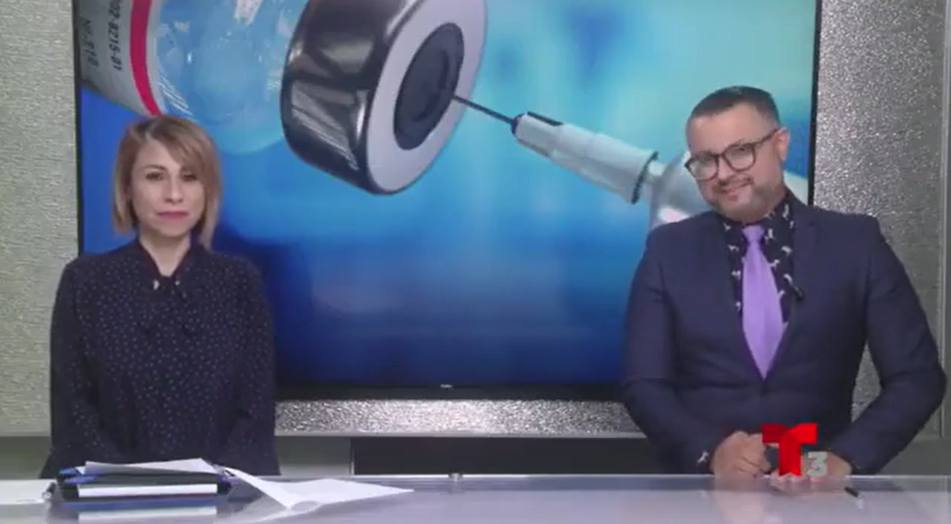 Two people sit down for a news interview in front of an image of a vaccination.