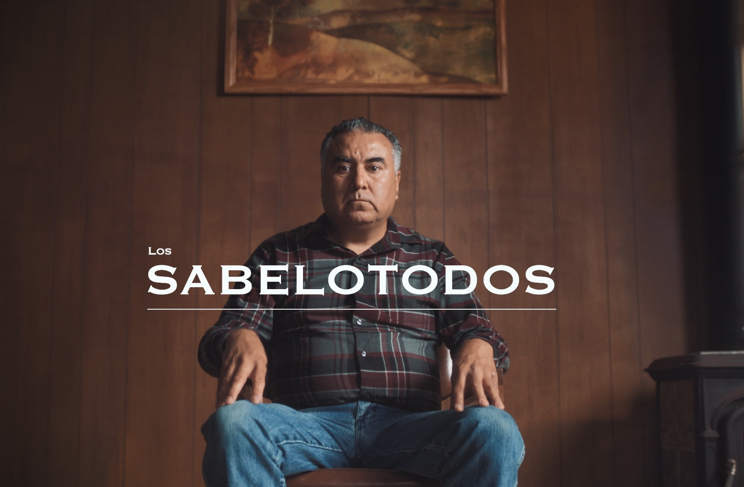 A Hispanic/Latino man sits in a chair in a wood-paneled room. The Spanish title reads, "The Know-It-Alls."