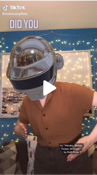 Screengrab of a Tiktok shows a young woman wearing a brown shirt and a space helmet. She appears to be doing the robot dance move 