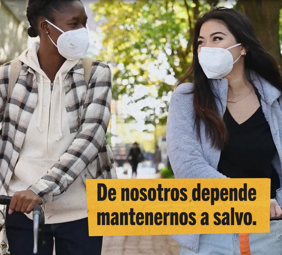 Two women of color walk together in the park. Both are wearing masks. Spanish text reads: "It's up to us to keep each other safe."