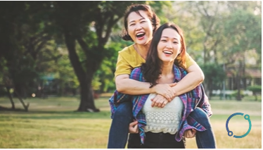 Image of a smiling Asian woman piggyback riding on a happy Asian woman in a park outside in the summer