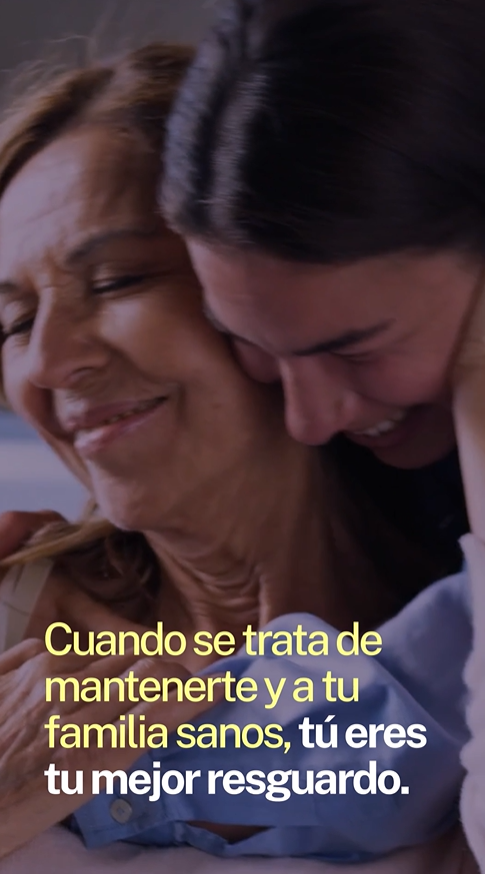 A granddaughter leans over her grandmother's shoulders and hugs her; both have big smiles on their faces.