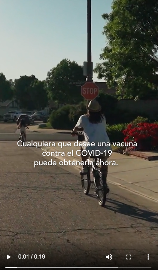 A child and an adult ride their bikes on the street with text overlaid noting that anyone can get a vaccine who wants one (specific eligibility criteria shared later in video)