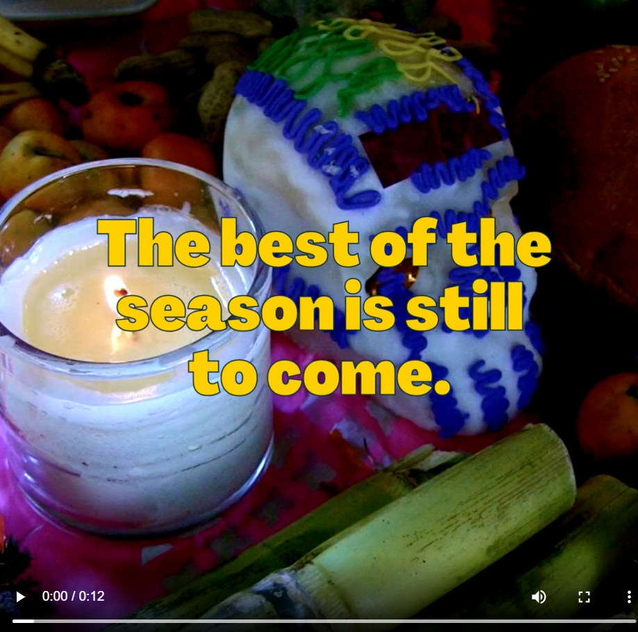 A lit candle and a colorful skull sit on a table. The words "the best of the season is still to come" appears in yellow