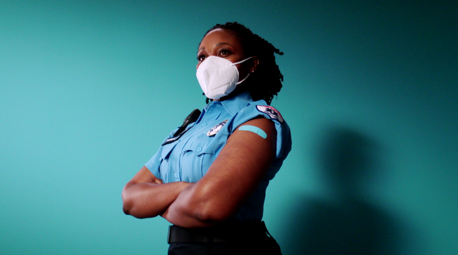 A Black woman wearing a mask crosses her arms and is wearing a band aid on her shoulder