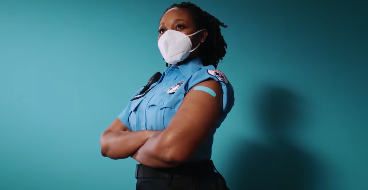 Black female police officer in uniform who is wearing a mask and has a band-aid over her vaccination site on her upper left shoulder.