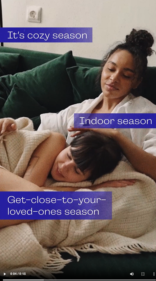 A young Black woman sits on the couch wrapped in blankets with her girlfriend. Text reads, "It's cozy season. Indoor season. Get-close-to-your-loved-ones season. Protect each other. Wash your hands, rock your mask, get your updated flu and updated COVID-19 shots."