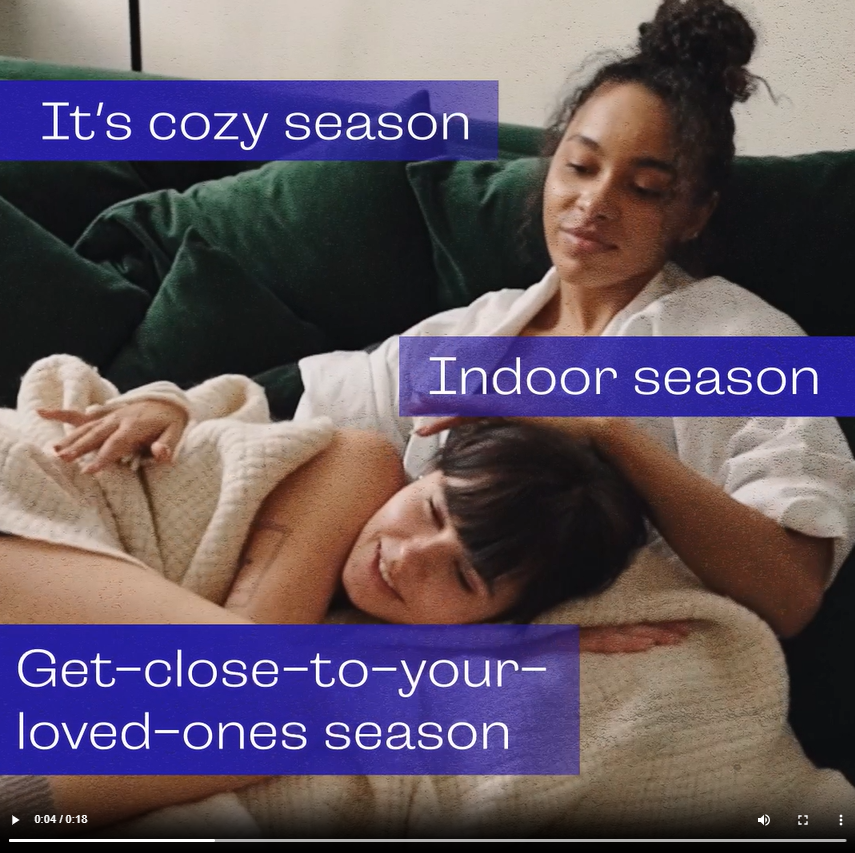 A young Black woman sits on the couch wrapped in blankets with her girlfriend. Text reads, "It's cozy season. Indoor season. Get-close-to-your-loved-ones season. Protect each other. Wash your hands, rock your mask, get your updated flu and updated COVID-19 shots."
