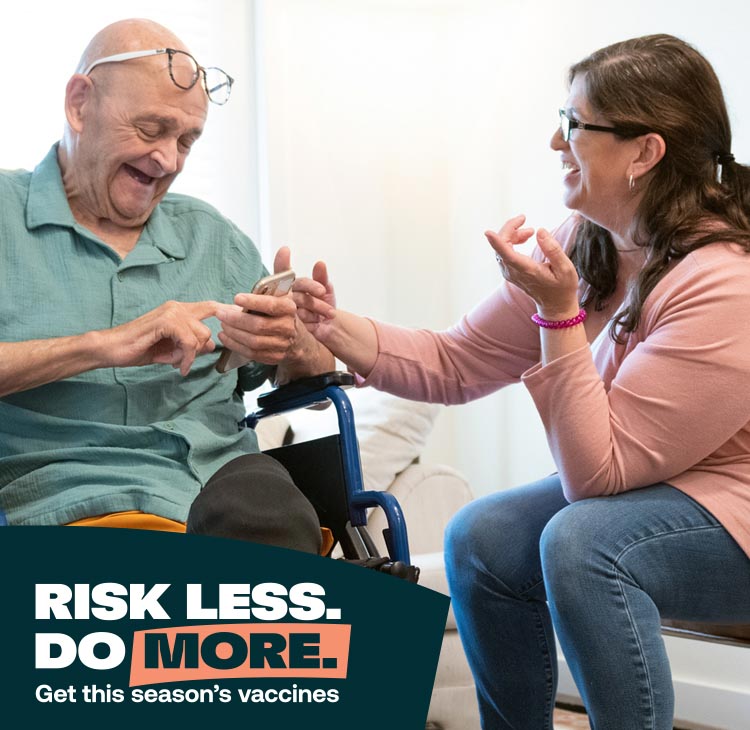 An older man in a wheelchair laughs while looking at his phone, next to a younger woman who's smiling.