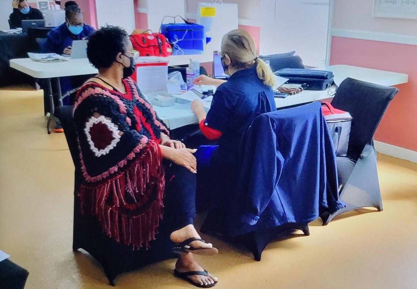 A masked person sits at a vaccination station while a nurse prepares to administer a vaccine