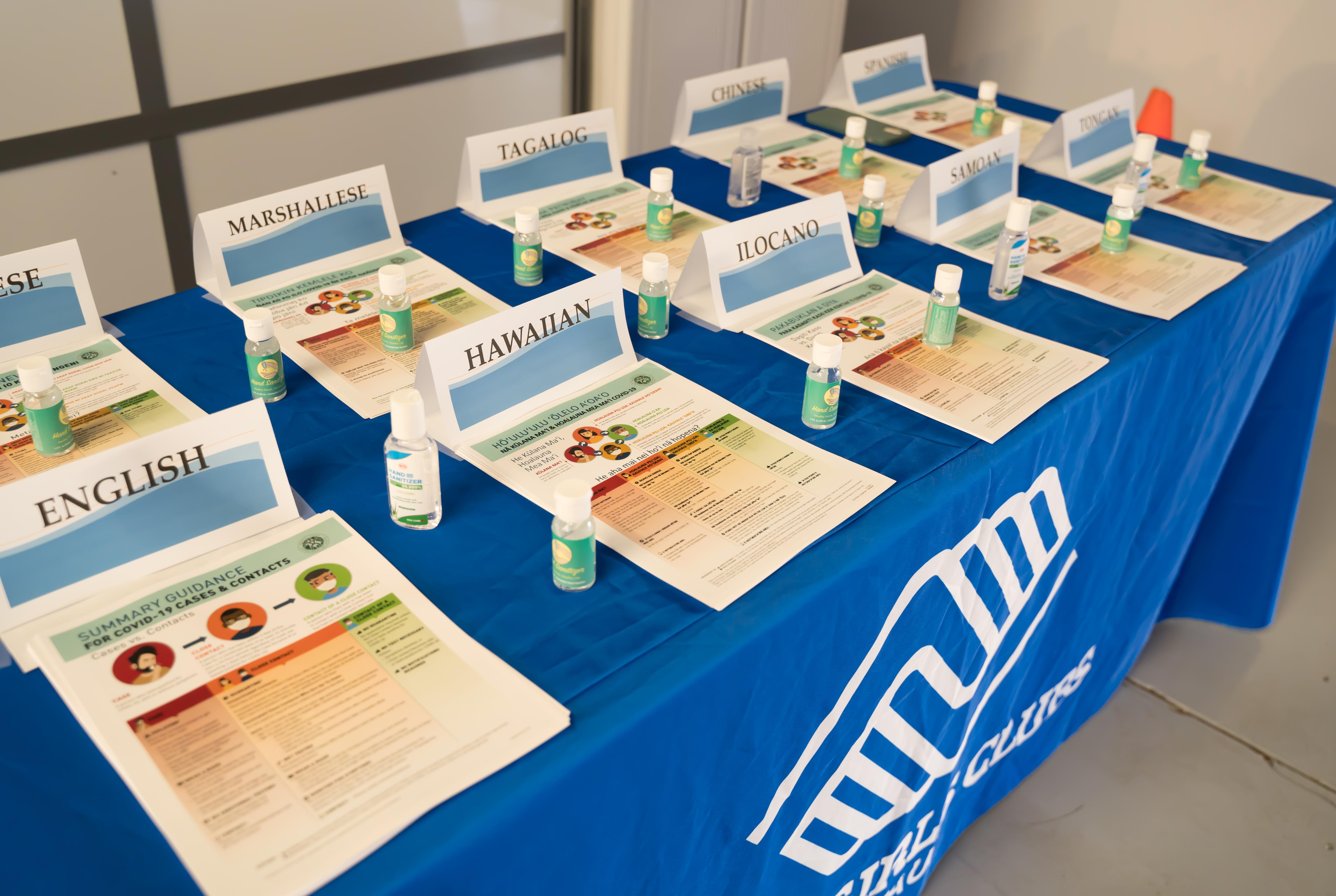 A display table contains numerous bottles of hand sanitizer and stacks of informational flyers with multiple languages available