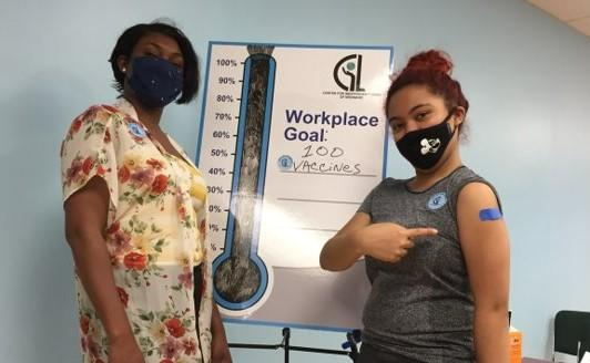 Two people wearing masks pose in front of a vaccine tracker poster while one shows a vaccine bandage on their arm