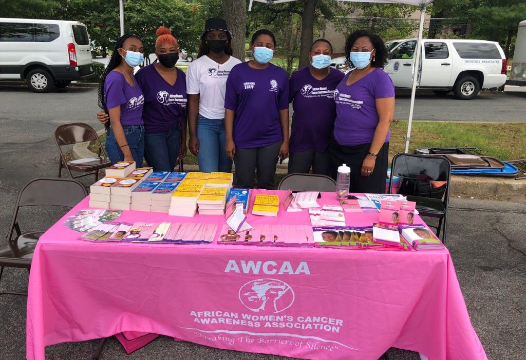A group of masked volunteers pose together behind a table that contains literature and books at an outdoor event,