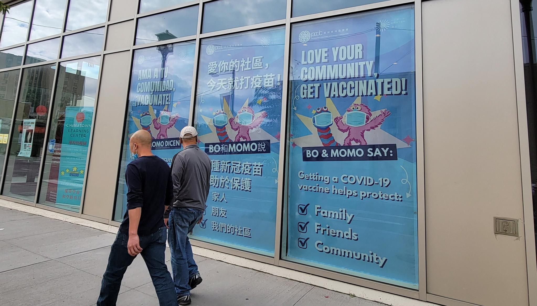 Two men walk past windows that display signs that say "love your community, get vaccinated!"