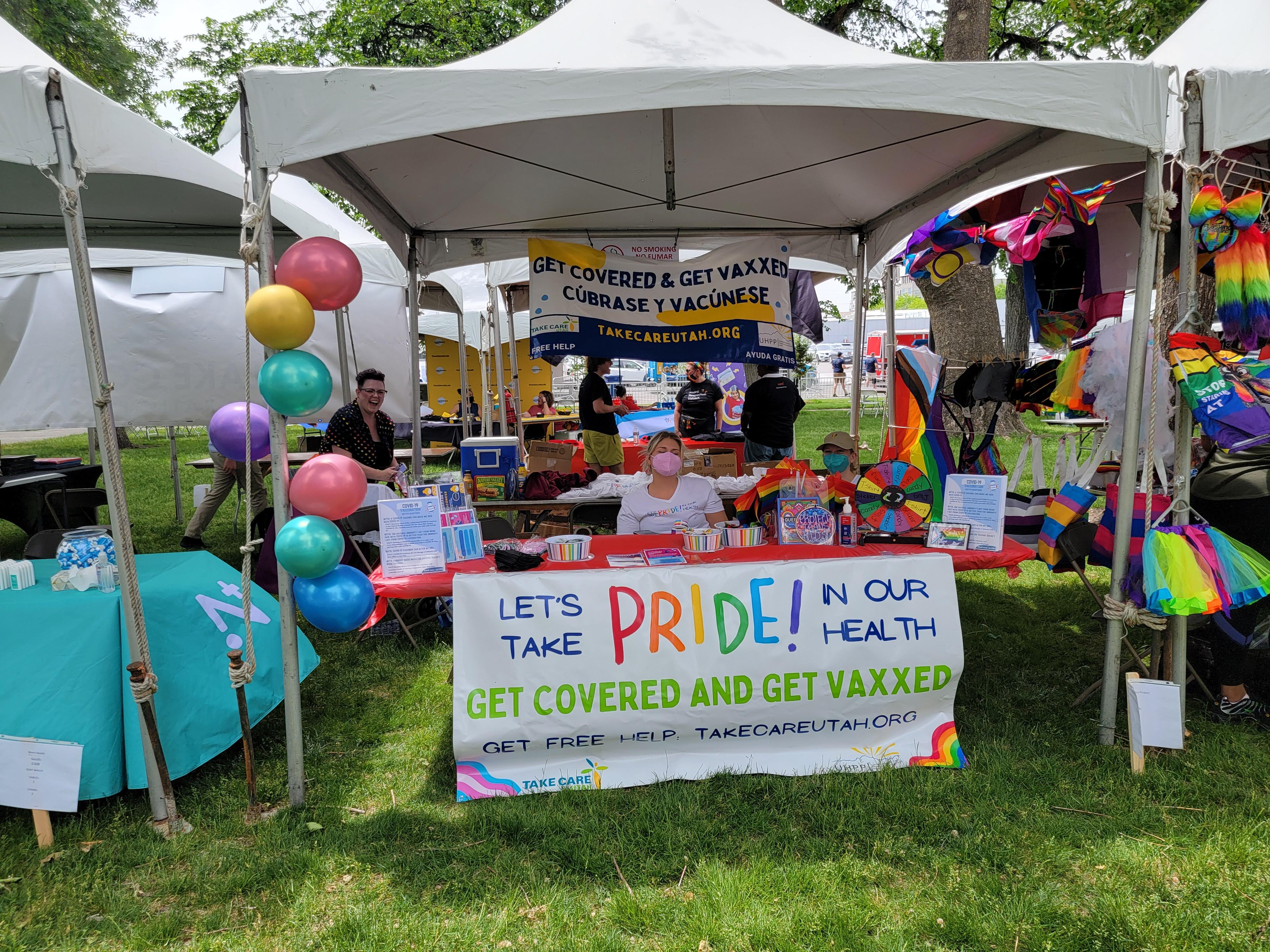 A woman wearing a mask sits at a table and welcomes people to the pride-themed vaccination event