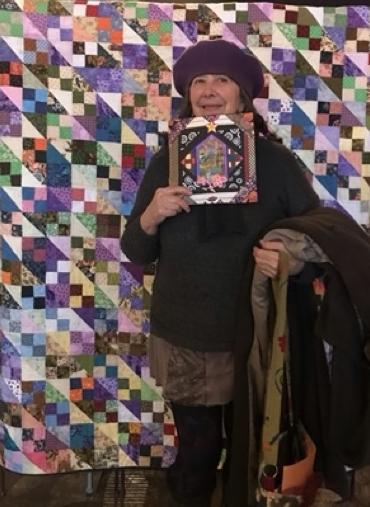 An Asian woman poses in front of a colorful barn quilt. 