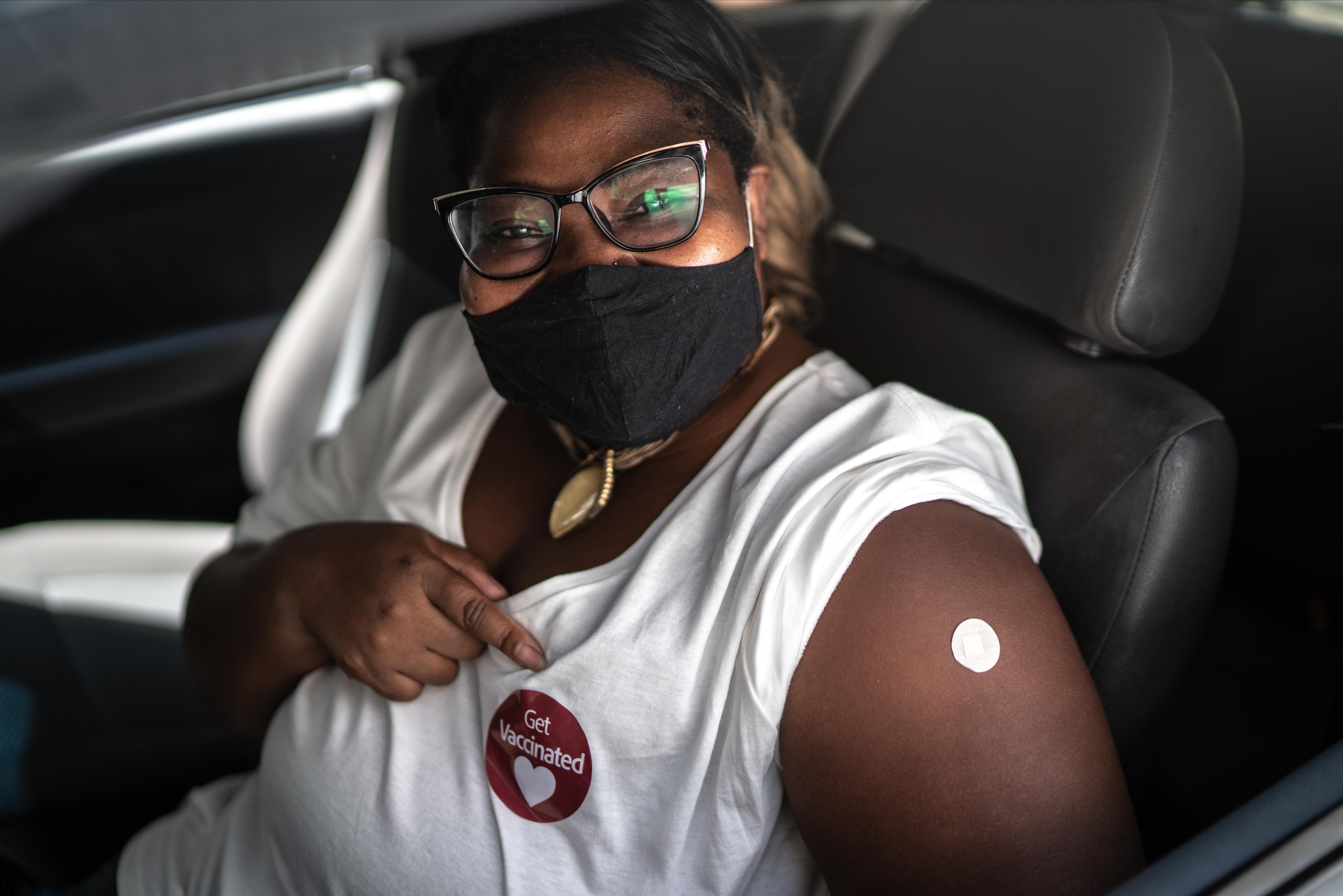 A Black woman wears a face mask and points to a sticker reading "Get Vaccinated!" on her chest. She has an adhesive bandage on her shoulder.
